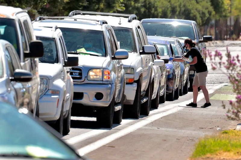 line of cars with masked person approaching a driver's window