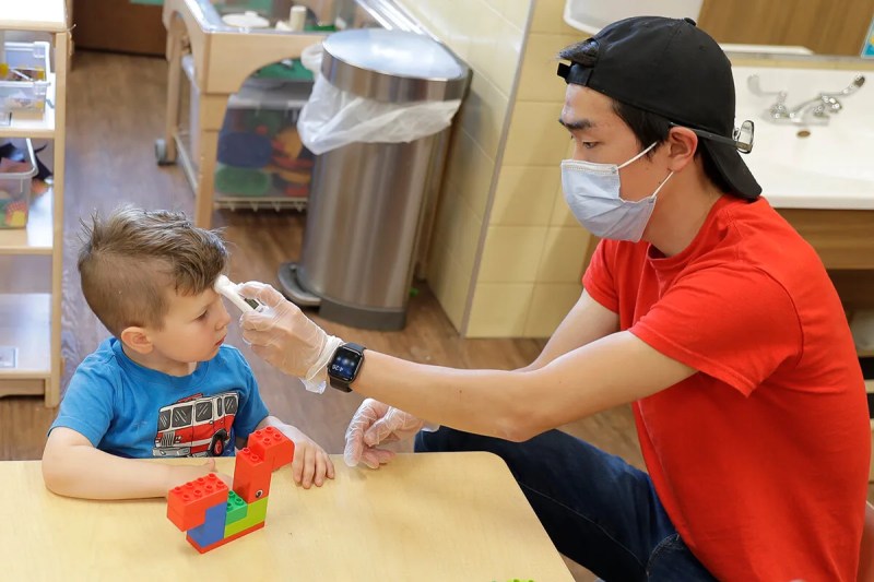 man holds thermometer up to young boy's forehead at childcare center