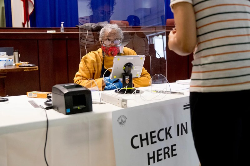 older poll workers sits behind plastic shield at check-in table wearing protective equipment