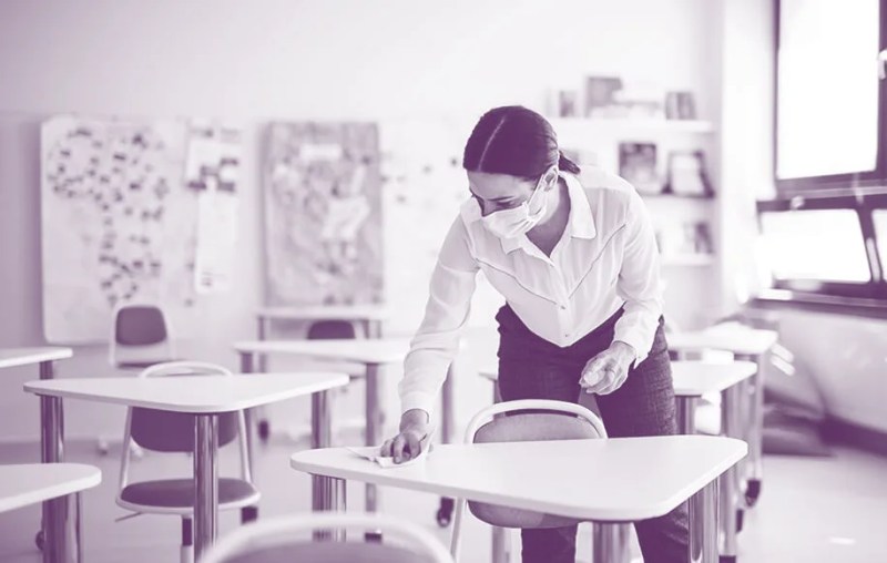woman wearing face masks cleans a desk in a classroom