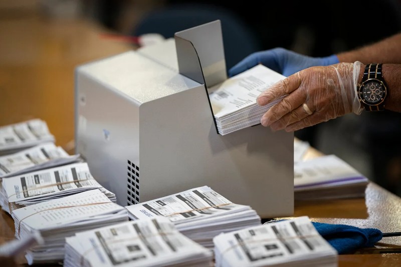 stacks of ballots next to hand placing ballots in processing machine