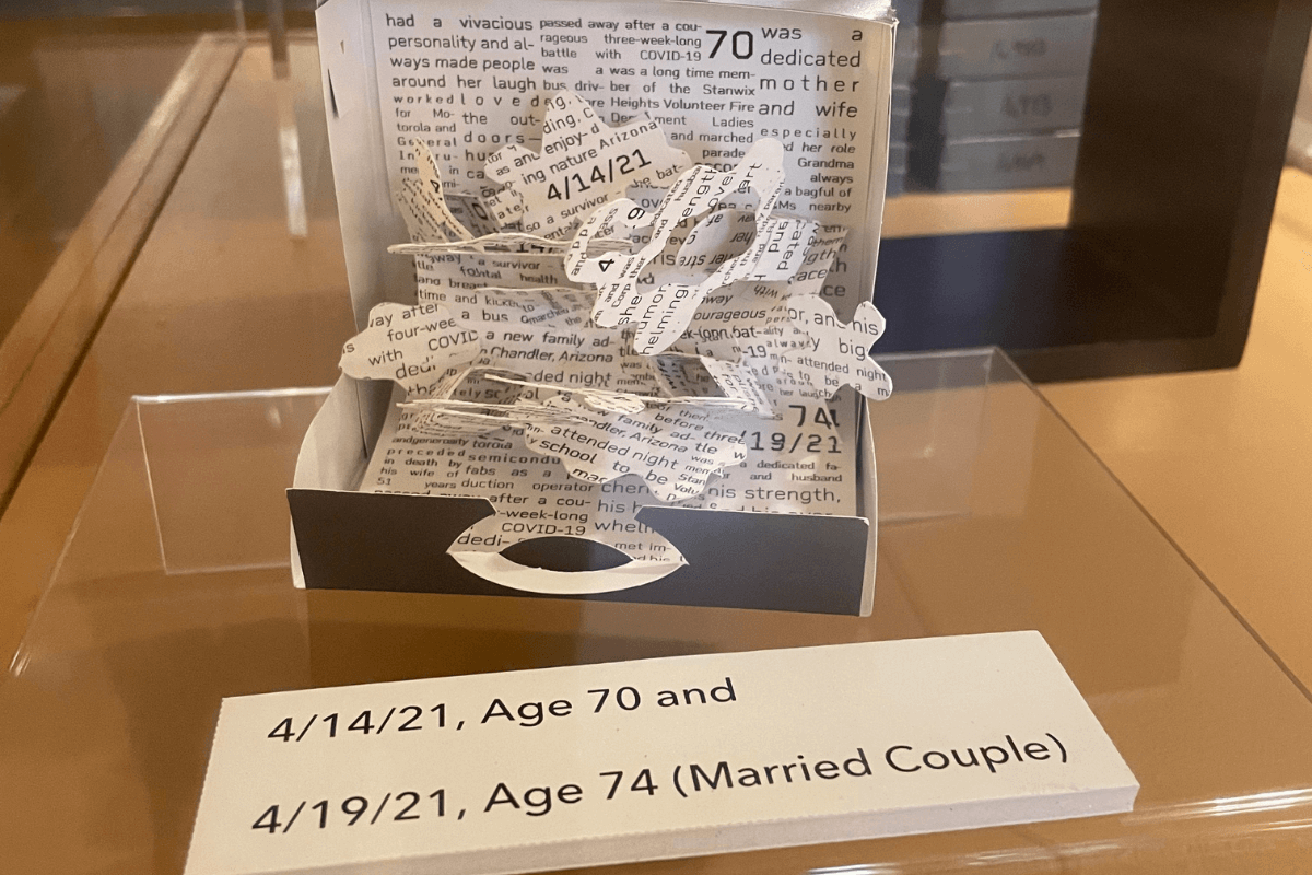 paper suitcase with origami inside made from an obituary. sign in front reads "4/14/21 age 70 and 4/19/21 age 74, married couple