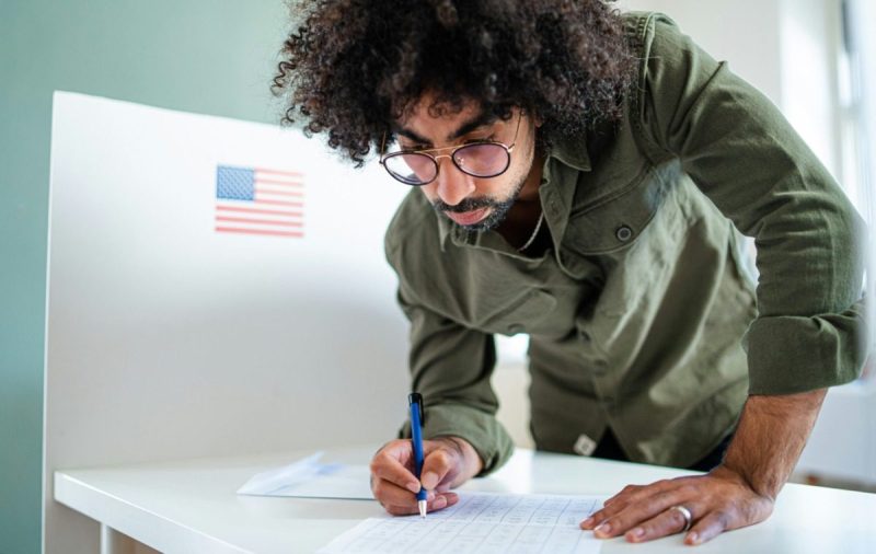 Young black man wearing glasses, filling out a voter ballot.