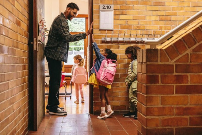 Elementary school teacher greeting his students at the door. Male teacher welcoming his class with a high five outside their classroom. Child mentor motivates his pupils in a primary school.
