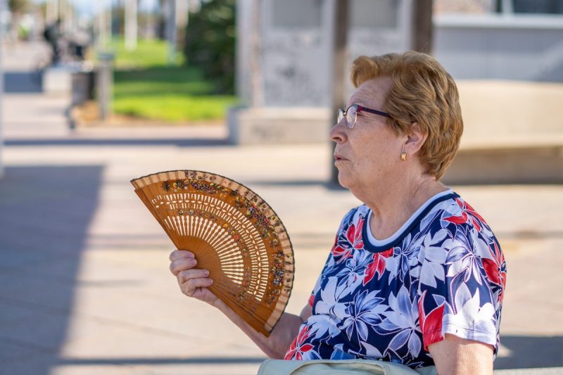 An older woman fanning herself on a hot day, sitting on a bench in the city.