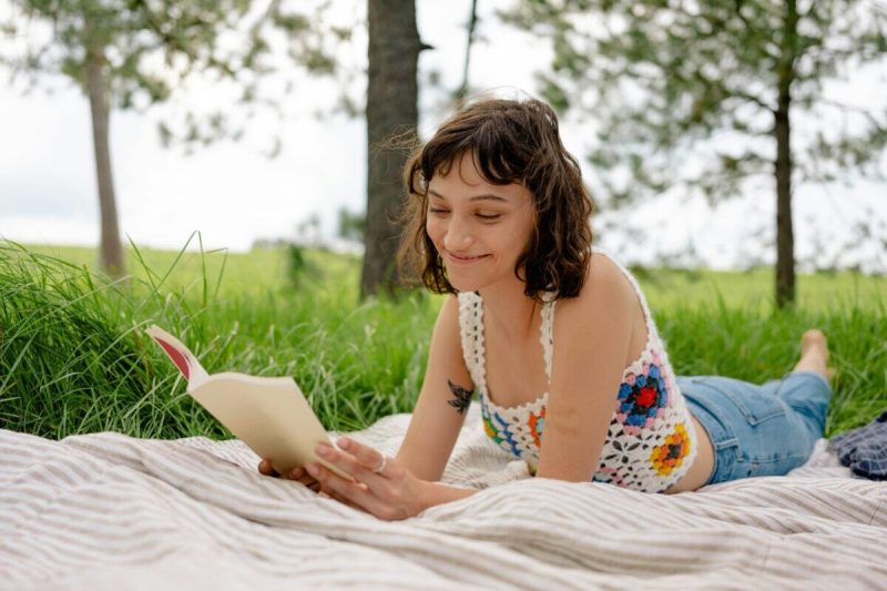 a woman lies on her stomach on a blanket outside, reading a book