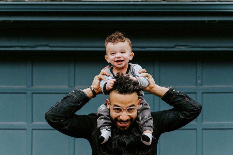 man holding infant on his shoulders in front of a teal garage door