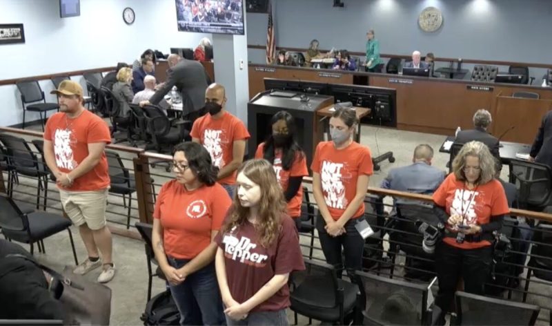people wearing red shirts standing in a government room with their backs to the panel sitting up front