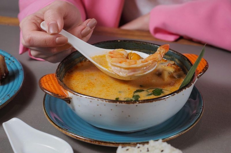 someone holding up a spoon with a shrimp on it above a bowl or orange soup