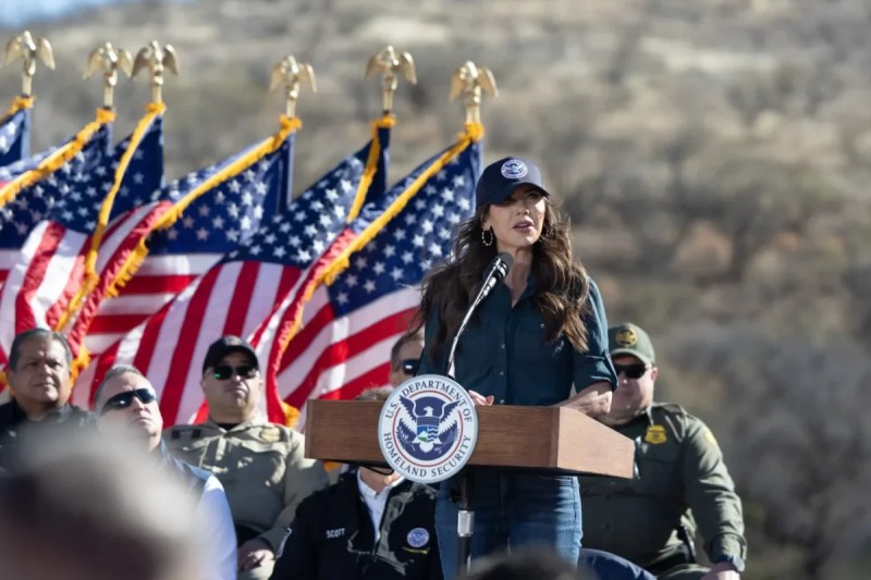 Kristi Noem talking at podium with a bunch of American flags flying behind her