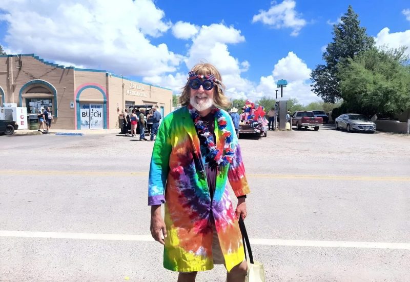 man standing on street wearing tie dye, a blonde wig, and American flag glasses